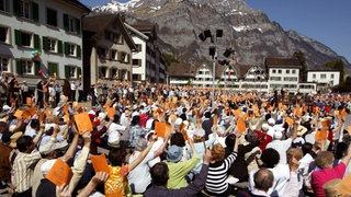 Auf einem Marktplatz, umstanden mit alten Häusern, im Hintergrund Berge, sitzen viele Menschen auf Bänken und heben orangefarbene Karten in die Höhe.