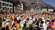 Auf einem Marktplatz, umstanden mit alten Häusern, im Hintergrund Berge, sitzen viele Menschen auf Bänken und heben orangefarbene Karten in die Höhe.