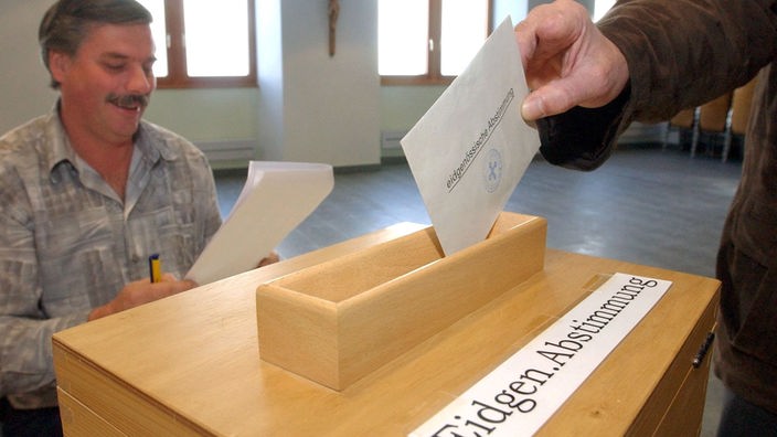 Ein Mann wirft einen Umschlag mit der Aufschrift 'eidgenössische Abstimmung' in eine Urne.