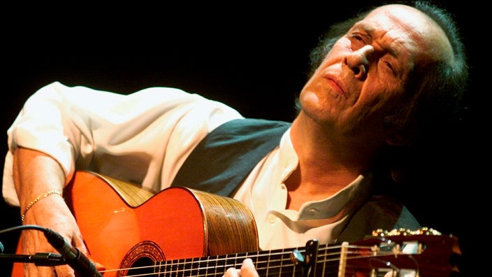 Paco de Lucia sitzt in weißem Hemd und schwarzer Weste auf einer Bühne und spielt Gitarre.