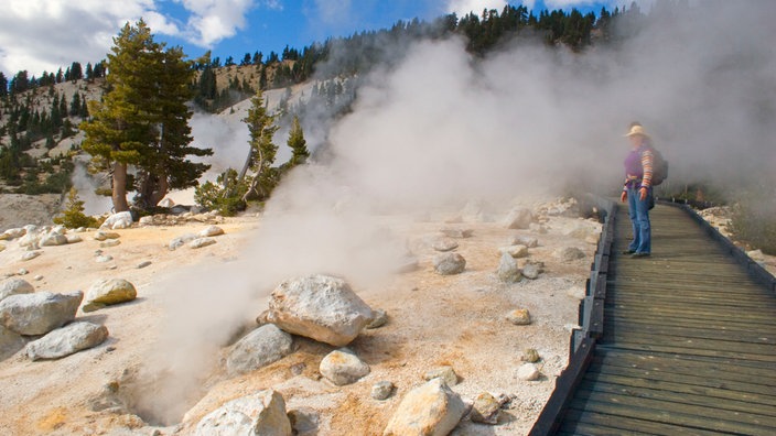 Ein Besucher des Lassen-Volcanic-Nationalparks steht auf einem hölzernen Pfad neben einem Erdloch aus dem heißer Dampf empor quillt. | Bildquelle: picture-alliance / united-archiv Ein Besucher des Lassen-Volcanic-Nationalparks steht auf einem hölzernen Pfad neben einem Erdloch aus dem heißer Dampf empor quillt.
