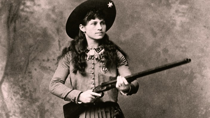 Das Schwarzweiß-Foto zeigt Annie Oakley stehend mit Cowboyhut, in einem mit Blumen besticktem Kleid und mit einem Gewehr in den Händen.