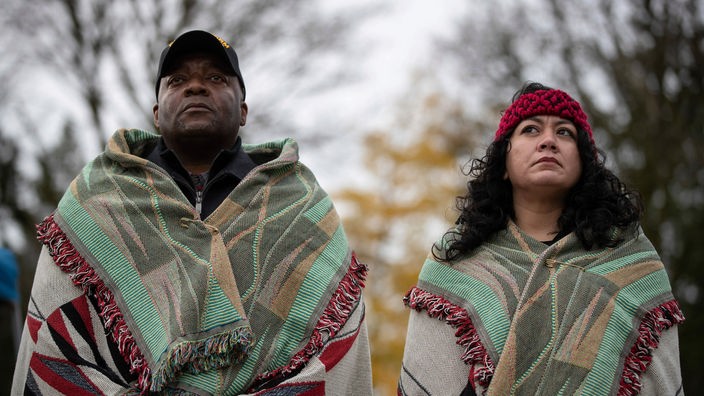 Ein schwarzer Mann und eine indigene Frau stehen nebeneinander und haben sich Decken über die Schultern gehängt