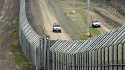 Weiter Blick über den Grenzzaun zwischen Mexiko und den USA. Auf der amerikanischen Seite patrouillieren US-Fahrzeuge.