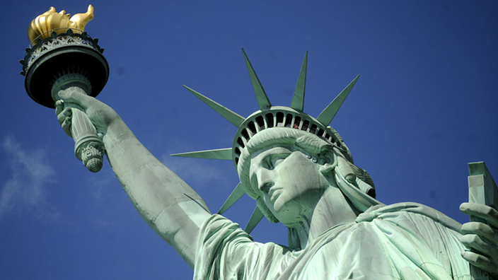 Zu sehen ist der Oberkörper der amerikanischen Freiheitsstatue vor blauem Himmel.
