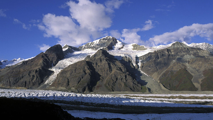 Das Bild zeigt einen weiß leuchtenden Gletscher auf dunkelgrauen, felsigen Bergen.