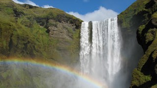 Von einem Felsen stürzt ein Wasserfall in die Tiefe. Ein Regenbogen liegt über dem fallenden Wasser.