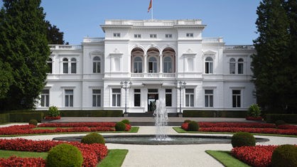 Das Foto zeigt die Villa Hammerschmidt. Vor dem prächtigen weißen Villengebäude ist ein Ziergarten angelegt, in dessen Mitte ein runder Springbrunnen steht. Auf dem Gebäude weht die Deutschland-Fahne.
