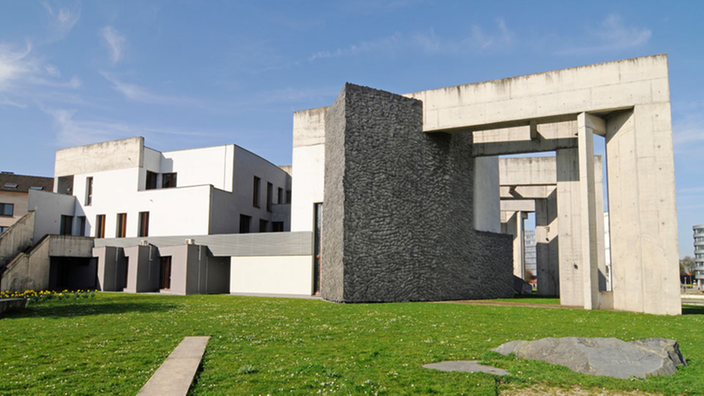 Auf einem Rasenstück steht ein halbrund angelegtes Gebäude. In regelmäßigen Abständen ragen graue Betonwinkel aus der Fassade.