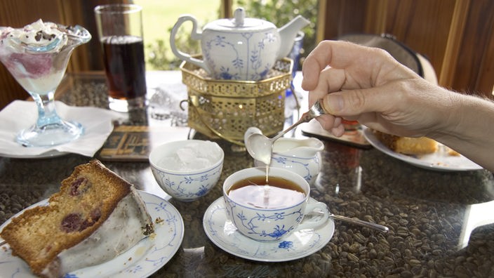 Ein Tisch ist für eine ostfriesische Teerunde gedeckt. Eine Hand lässt Sahne in eine schon mit Tee gefüllte Tasse tropfen. Daneben steht ein Teller mit einem Stück Kuchen und eine Teekanne mit filigranem Blumenmuster.