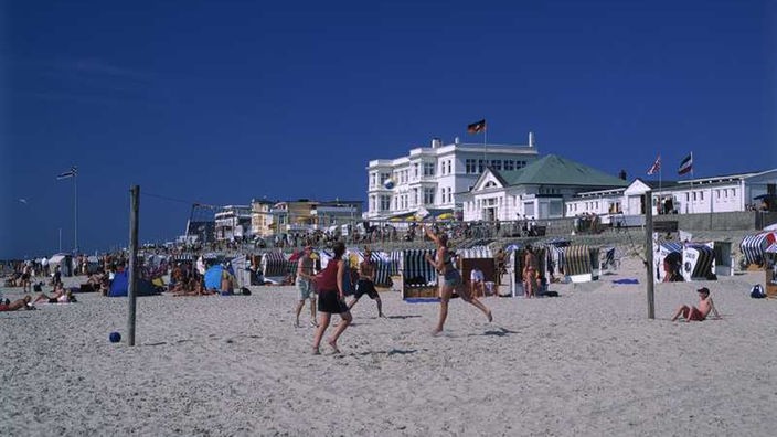 Das Bild zeigt eine Gruppe junger Leute am Strand, die Beachvolleyball spielen. Im Hintergrund ist die Strandpromenade zu sehen, an der mehrere elegante Villen stehen.