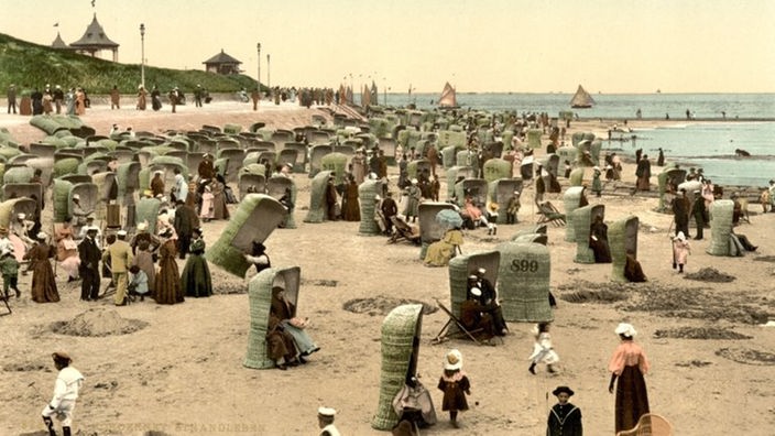 Das alte Foto zeigt den früheren Damenstrand Norderneys. Er ist voll gestellt mit alten Strandkörben aus Bast; es sind viele Frauen in langen Kleidern und Kinder mit Kleidern beziehungsweise Matrosenanzügen zu sehen.