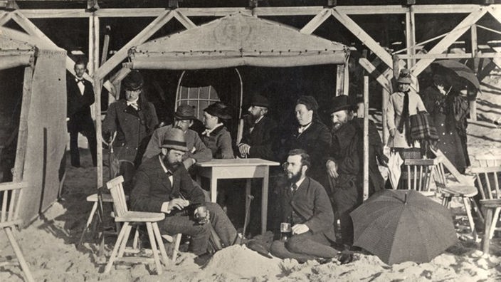 Schwarzweiß-Foto: feine Gesellschaft am Strand. Menschen in Anzügen und Kleidern sitzen unter einem Strandhaus um einen Tisch herum.