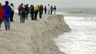 Menschen gehen im Winter am Strand spazieren und beobachten an einer Abbruchkante, wie das Meer den Sand fortspült.