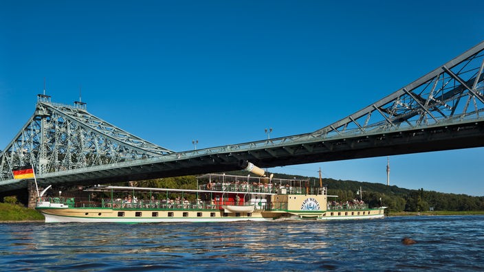 Das Bild zeigt ein Ausflugsboot auf einem Fluss, darüber erstreckt sich eine blau angestrichene Brücke