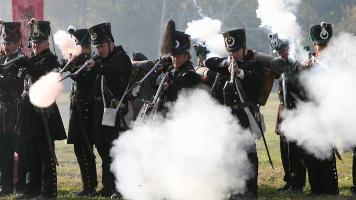 Nachgestellte Szene: Mehrere Männer in historischer Soldatenuniform geben Schüsse aus Gewehren ab.