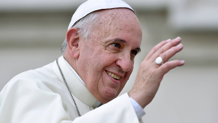 Papst Franziskus hebt lächelnd die Hand zum Gruß.