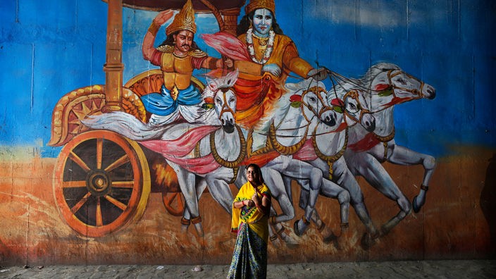 Eine Frau steht vor einem großen Wandgemälde mit einer Szene aus dem Mahabharata-Epos