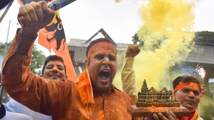 Ein schreiender Mann in Orange hält in einer Hand ein Tempel-Modell und in der anderen eine rauchende Fackel