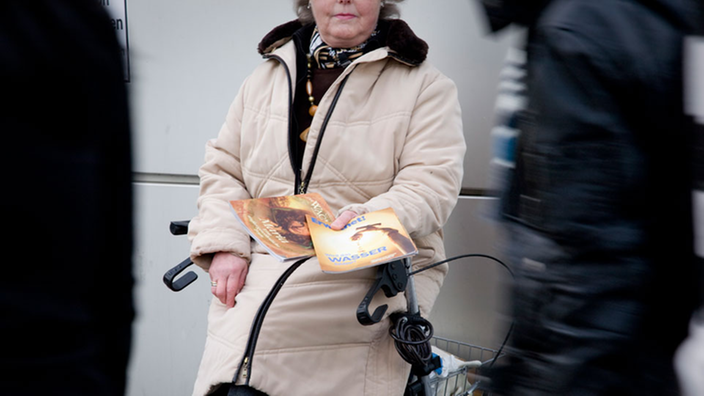 Eine alte Frau sitzt auf einem Schiebewägelchen in einer Fußgängerzone und hält zwei religiöse Zeitschriften in der Hand.