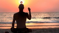 Ein Mann sitzt meditierend vor dem Sonnenuntergang an einem Strand und balanciert einen durchsichtigen Ball auf dem Kopf.
