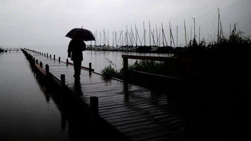 Eine Person mit Regenschirm steht allein auf einem Bohlensteg am Meer vor einem dunklen, wolkenverhangenen Himmel.