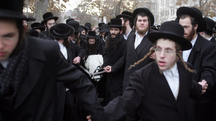 Mehrere orthodoxe Juden in schwarzer Kleidung, mit Hüten und Bärten. Einige von ihnen tragen einen Leichnam, der in ein weißes Tuch gehüllt ist.