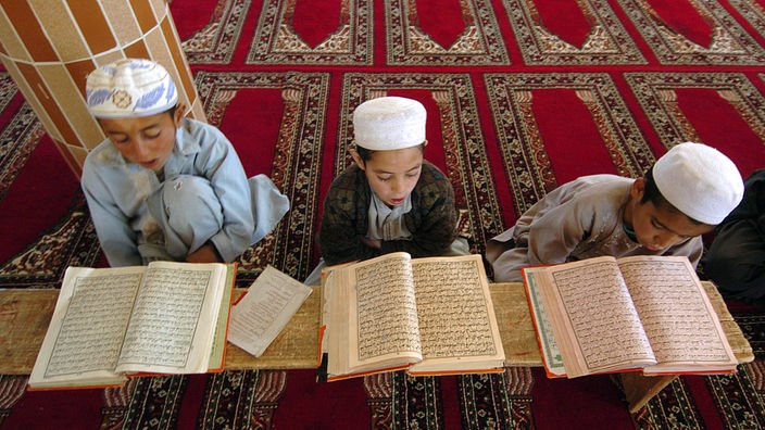 Drei muslimische Kinder sitzen auf Teppichen und lesen im Koran.
