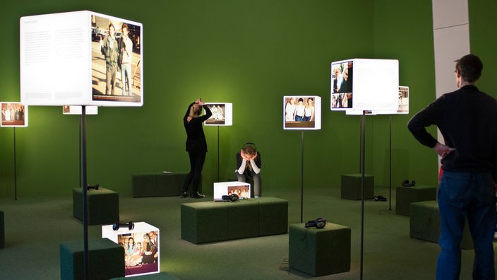Eine Führerin steht in einem Museumsraum mit mehreren Fotografien an der Wand und erklärt einigen Besuchern die Ausstellung.