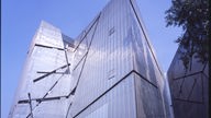 Das Jüdische Museum von außen. Zu sehen ist ein schief wirkendes, modernes Gebäude. Durch die metallisch glänzende Außenfassade ziehen sich Risse.