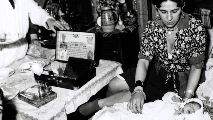Schwarzweiß-Foto: Eine Frau kümmert sich um ein Baby. Links sieht man einen Mann, der medizinisches Gerät aus einem Koffer nimmt.
