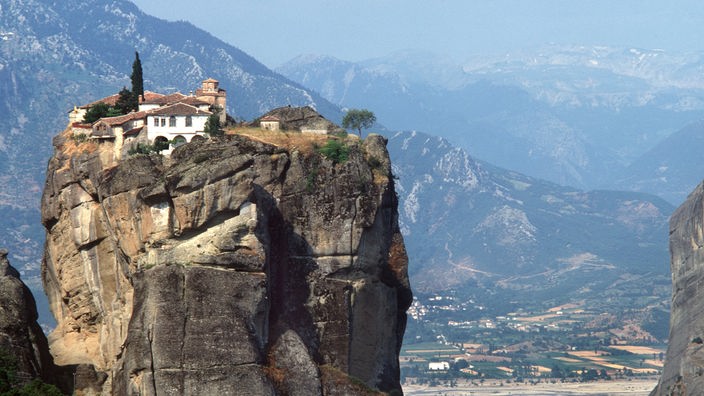 Auf einem hohen, schroffen Felsen stehen die Gebäude einer Klosteranlage. Im Hintergrund sind Berge zu erkennen.