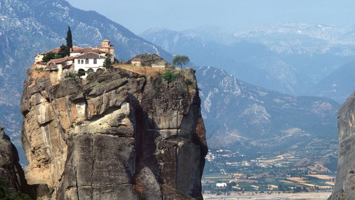 Auf einem hohen, schroffen Felsen stehen die Gebäude einer Klosteranlage. Im Hintergrund sind Berge zu erkennen.