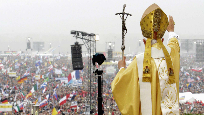 Der Papst steht erhöht vor einer riesigen Menschenmenge und hat den rechten Arm zum Segen ausgestreckt.