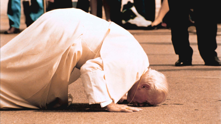 Der Papst in weißer Kluft kniet auf dem Boden, hat sich nach vorn gebeugt und küsst die Erde.