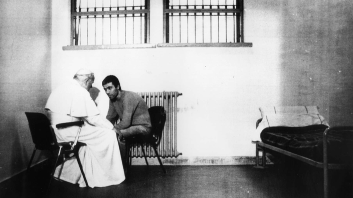 Das Schwarzweiß-Foto zeigt Papst Johannes Paul II. in weißer Kleidung. Er sitzt in einer Gefängnisstelle auf einem Stuhl. Ihm gegenüber sitzt ein jüngerer Mann, der sich zum Papst nach vorn beugt.