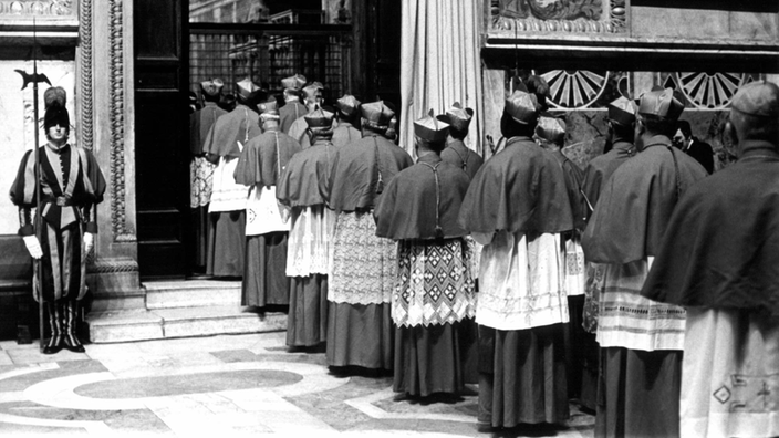 Das Schwarzweiß-Foto zeigt gut 20 Kardinäle, die in Zweierreihen stehen und durch eine große Tür in die Sixtinische Kapelle gehen.