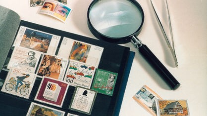 Briefmarken sind in eine Mappe einsortiert. Daneben liegen Lupe und Pinzette.  