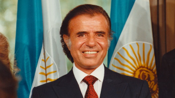 Carlos Menem lächelt in die Kamera. Im Hintergrund die argentinische Flagge.