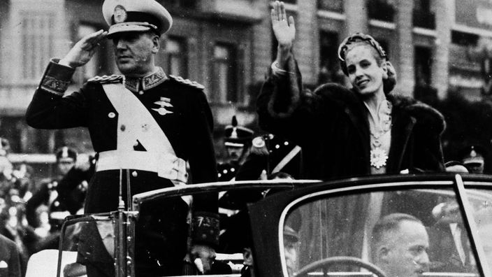 Perón und Evita in einem offenen Wagen bei einer Fahrt durch die Straßen. Er hat eine Uniform an und salutiert, sie trägt ein schickes Kostüm und winkt.
