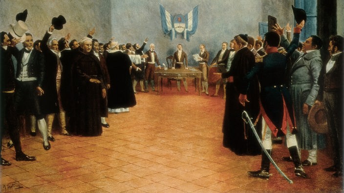Gemälde von der Versammlung in Tucumán. Rechts und links stehen Männer, die Hüte in die Luft halten und damit abstimmen. Im Hintergrund ein Pult, über dem die argentinische Nationalflagge hängt.