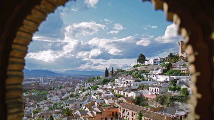 Durch einen Rundbogen in der Mauer der Alhambra von Granada kann man auf das gegenüber liegende Stadtviertel Albaicin blicken, dessen weiße Häuser an einem Hang liegen.