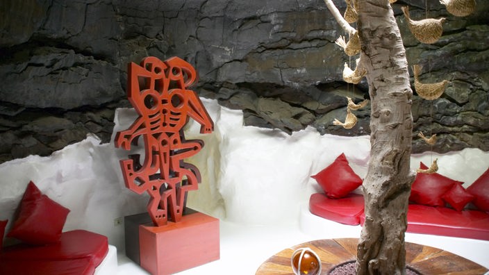 Ein unterirdischer Raum mit einer Bank mit Kissen und einer roten Skulptur. Am rechten Bildrand ist ein Baumstamm zu sehen. Die Wände sind aus Lavagestein.