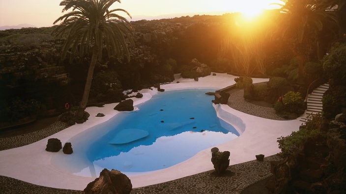Blick auf einen weißen Swimmingpool in organischer Form mit blauem Wasser bei Sonnenuntergang. Am Beckenrand stehen Palmen.