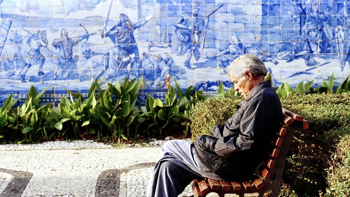 Mann sitzt auf Parkbank, dahinter Wand aus blauen Azulejos.