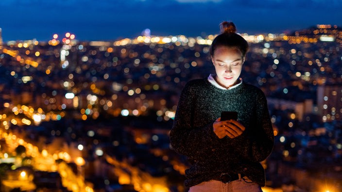 Eine junge Frau blickt auf ihr Smartphone, im Hintergrund das erleuchtete Barcelona