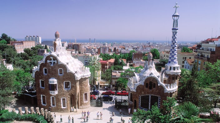 Blick von oben auf zwei reich verzierte Gebäude im 'Parc Güell' in Barcelona. Dahinter Panoramablick über das Häusermeer der Stadt, bis am Horizont das Meer zu sehen ist.