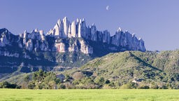 Blick über eine Wiese und bewaldete Hänge auf die bizarren Felsnadeln des Montserrats vor strahlend blauem Himmel.