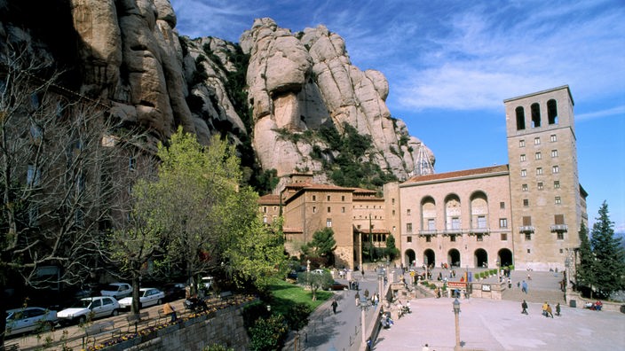 Im Hintergrund das Kloster Montserrat an einem Felsen. Von der linken Seite führt eine Straße vor das Kloster, die mit Autos zugeparkt ist. Rechts ein Platz mit zahlreichen Menschen.