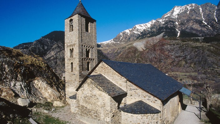 Blick auf eine alte kleine Kirche. Im Hintergrund felsige und verschneite Berge.